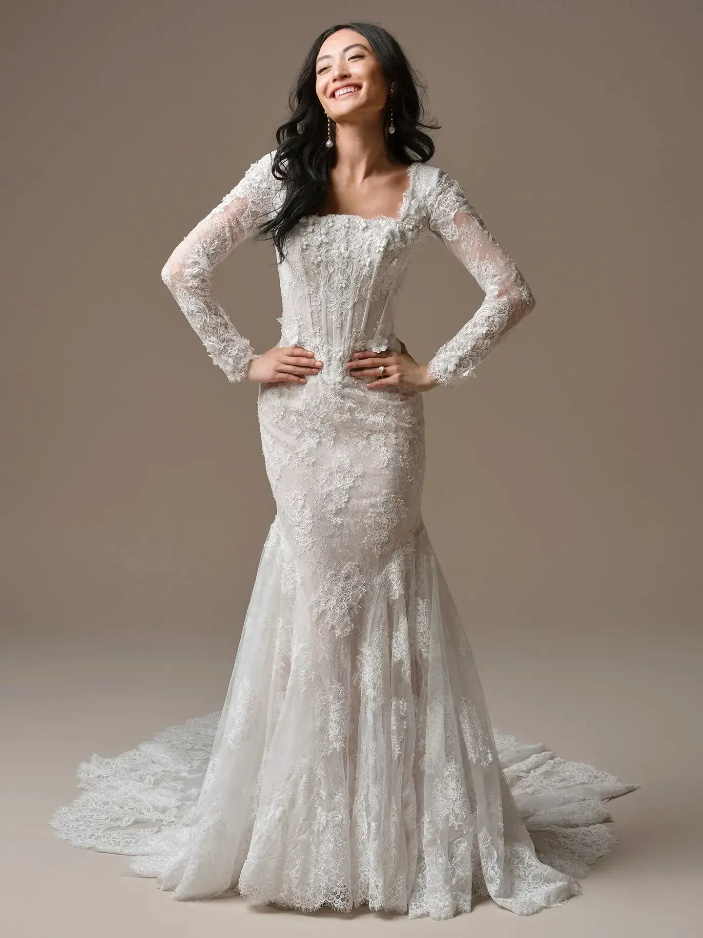 A woman in a lace wedding dress with long sleeves stands confidently, smiling. The dress features a fitted bodice and a flowing train.