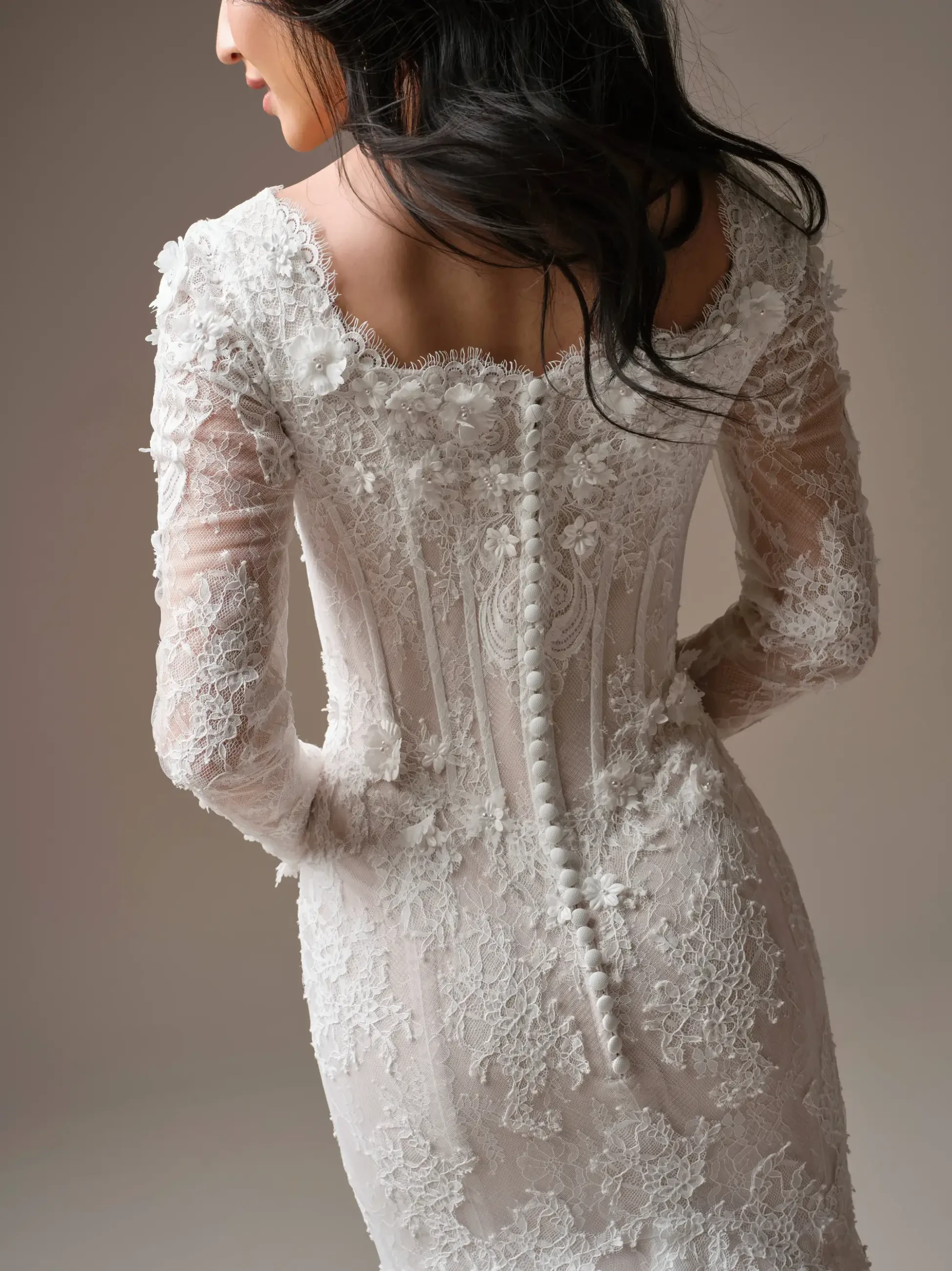 A bride stands in an elegant lace wedding gown, facing away. The dress features intricate floral patterns and delicate buttons, conveying a sense of romance.