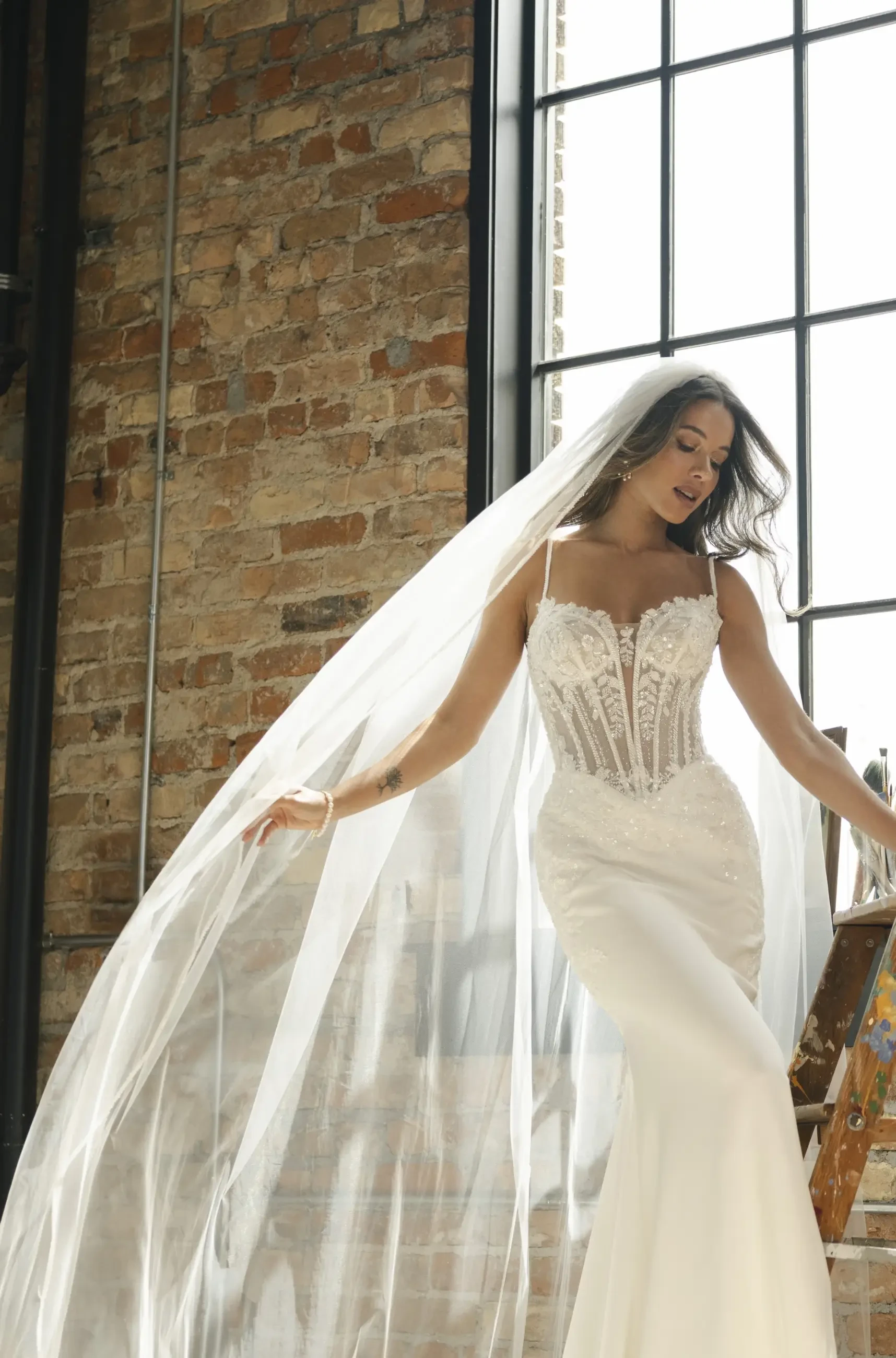 Bride in elegant white gown with lace bodice and flowing veil stands near rustic brick wall and large window, exuding elegance and serenity.