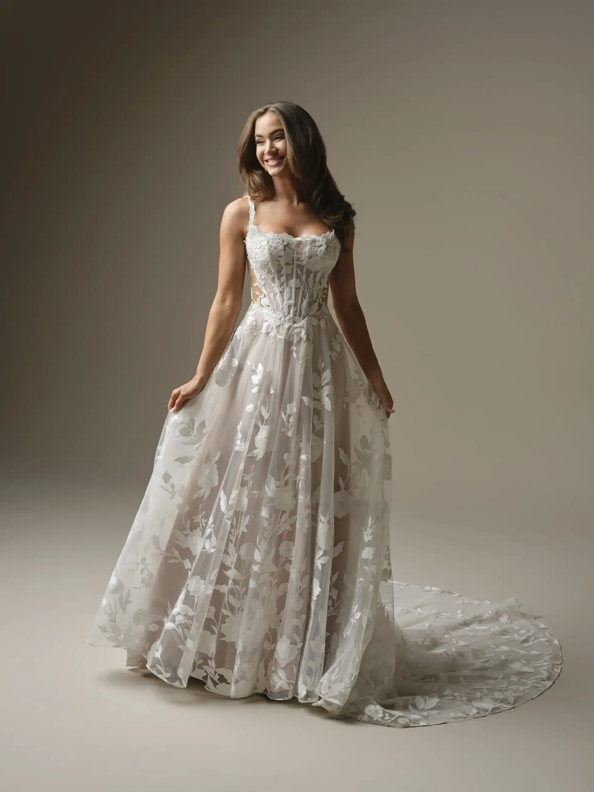 A woman in a beautiful, floral-patterned wedding dress with a flowing skirt and a train, standing against a neutral background.