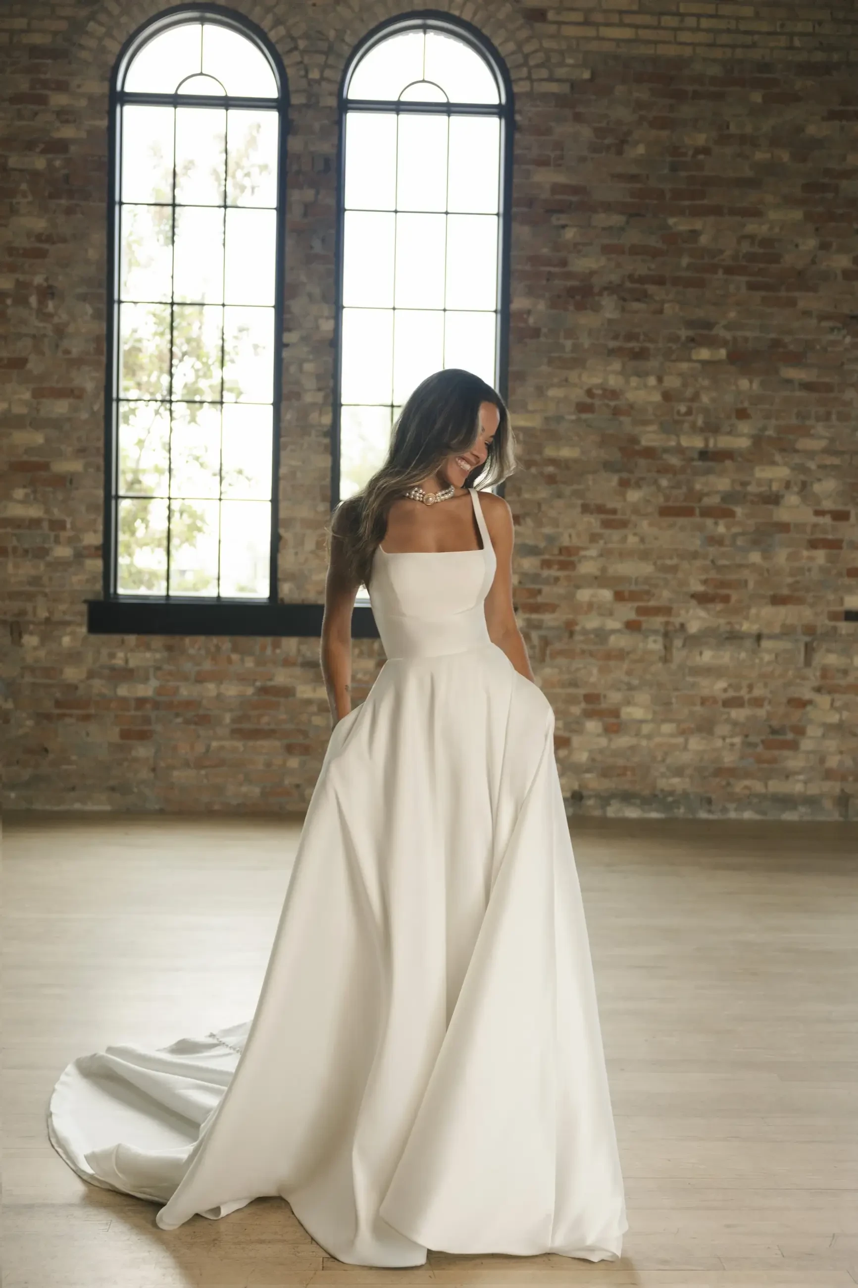 A woman in a white dress standing in a room with large windows and brick walls.