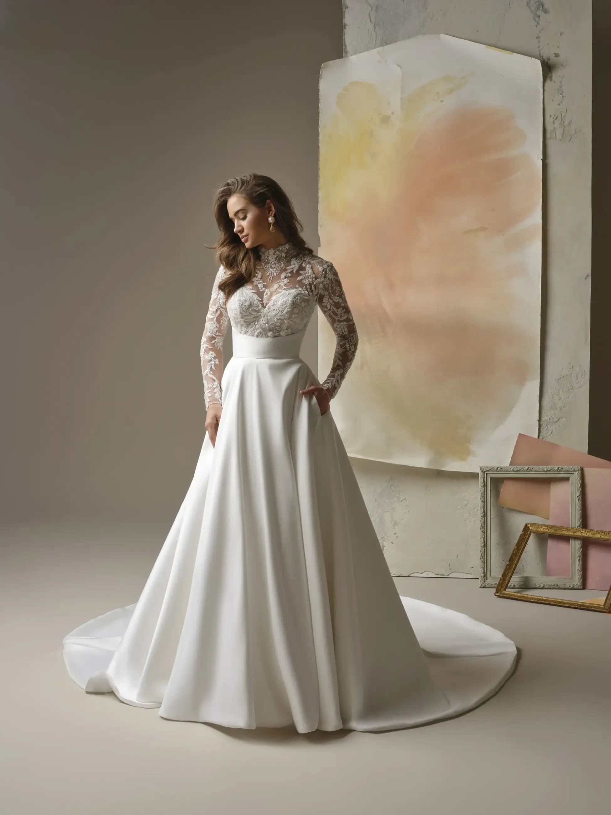 A bride in an elegant, long-sleeved lace wedding gown with a satin skirt, stands in a softly lit room. The ambiance is serene and sophisticated.