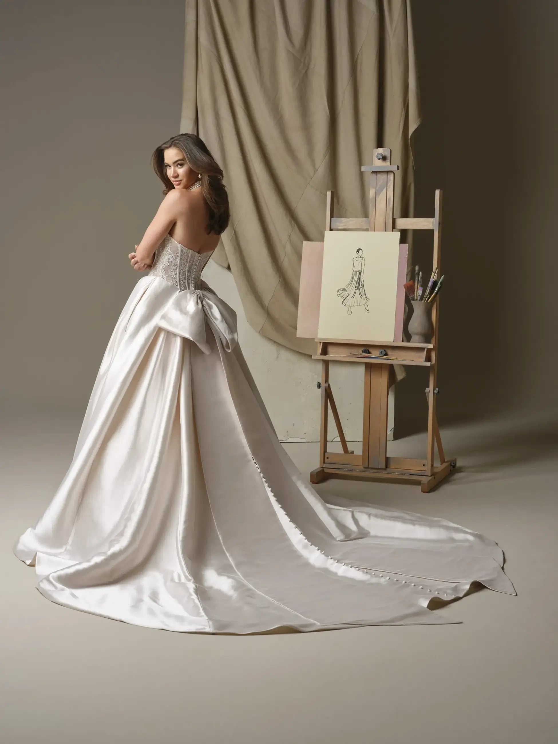 A woman wearing a white gown with a flowing train stands gracefully, facing away from the camera. She is positioned in front of an easel with a sketch of a dress. The background features draped fabric, creating an elegant atmosphere.