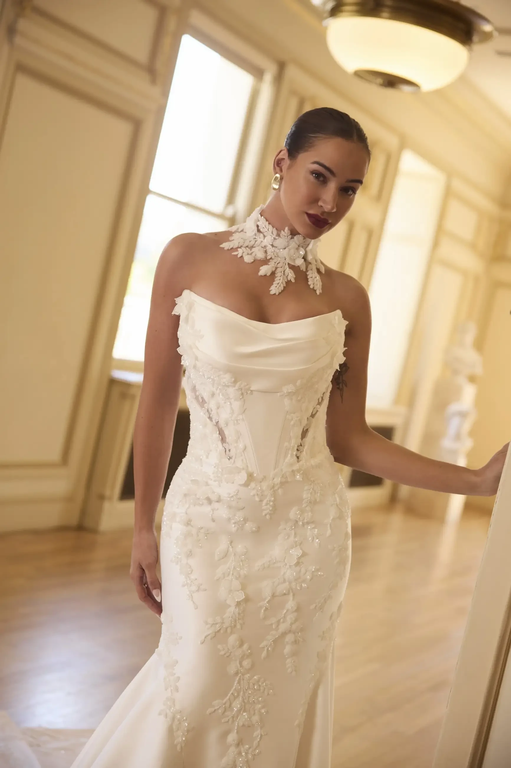 A woman in an elegant white gown with floral details poses in a beautifully lit room.