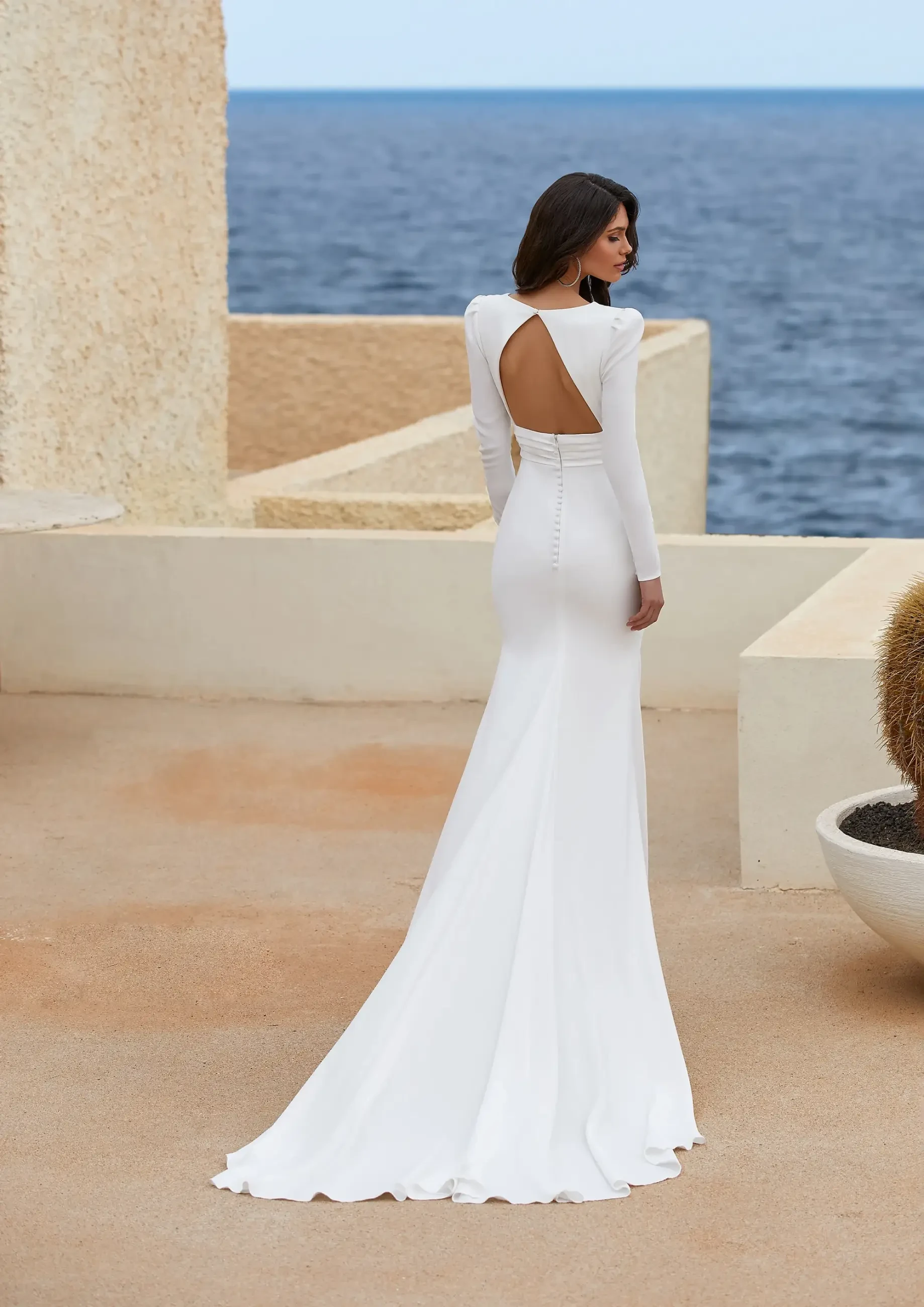 A woman in an elegant, long-sleeved white gown with an open back stands on a terrace overlooking the ocean. The scene conveys a serene and sophisticated mood.